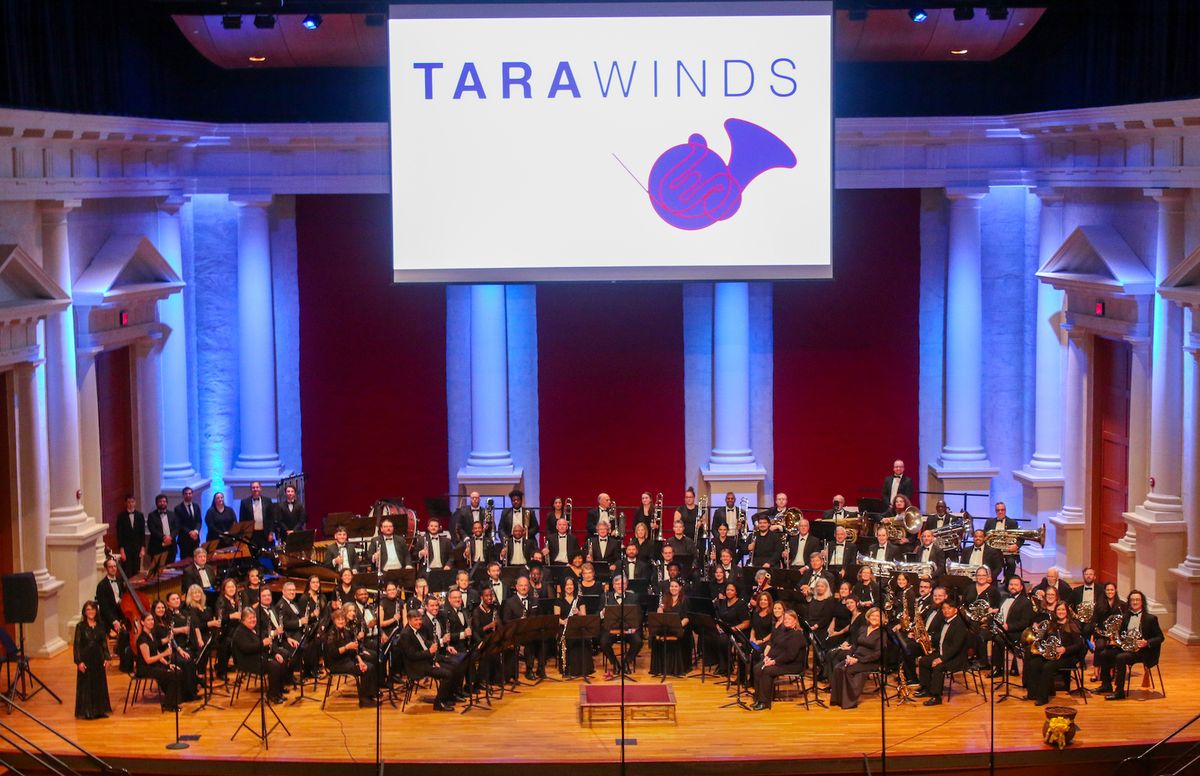 The full band in concert black on a columned stage beneath a projected Tara Winds mark